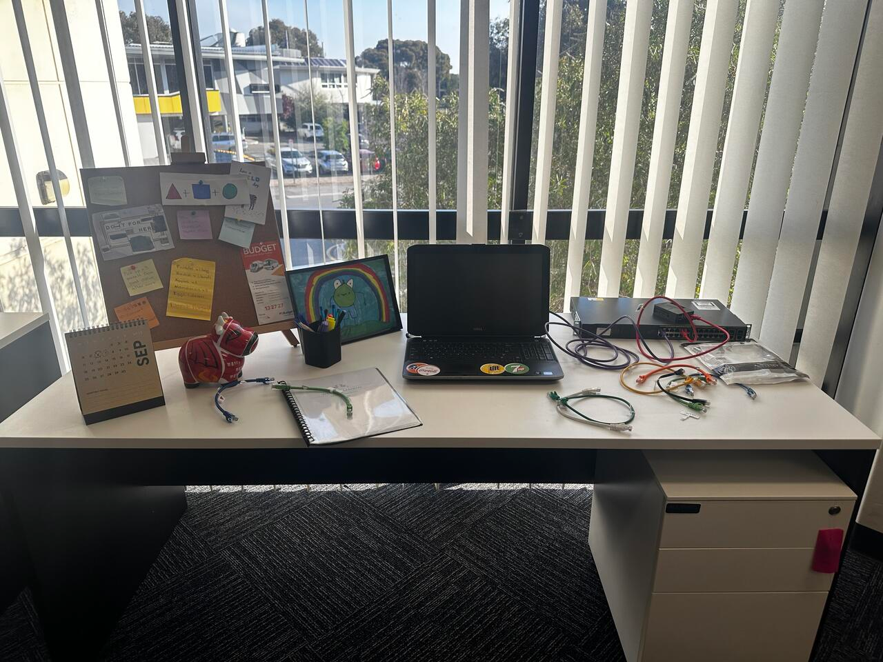 The network engineer's desk: a laptop, a switch, patch leads, a pinboard and a calendar.